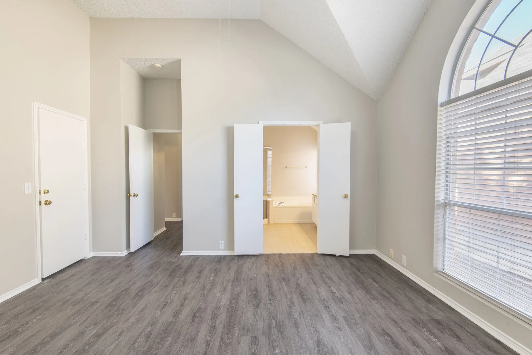 Vaulted ceiling, wood-finish flooring, arched window with horizontal blinds, neutral wall paint, and double-door entryway