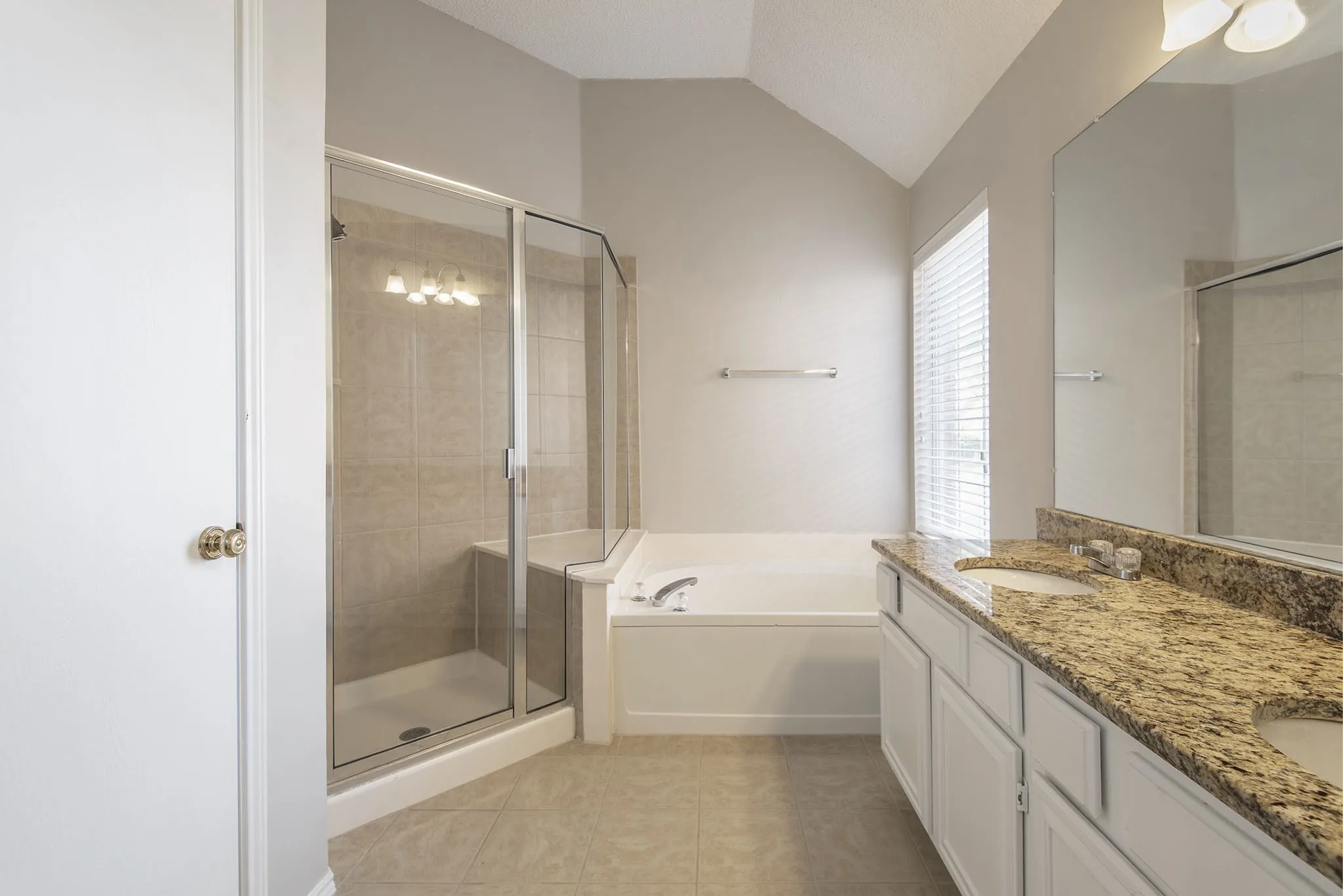 Vaulted ceiling, walk-in shower with tile surround, built-in soaking tub, dual vanity with granite-finish countertop, and tile flooring