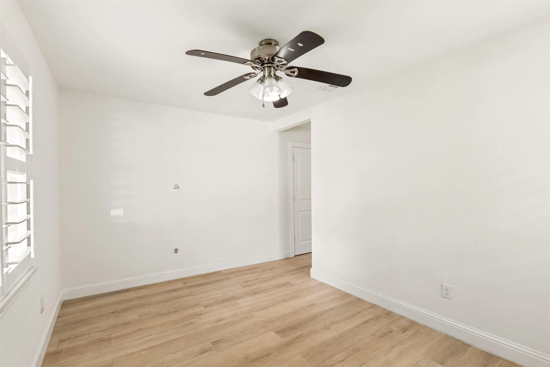 Wood-finish flooring, white walls, ceiling fan with integrated lighting, and window with plantation shutters
