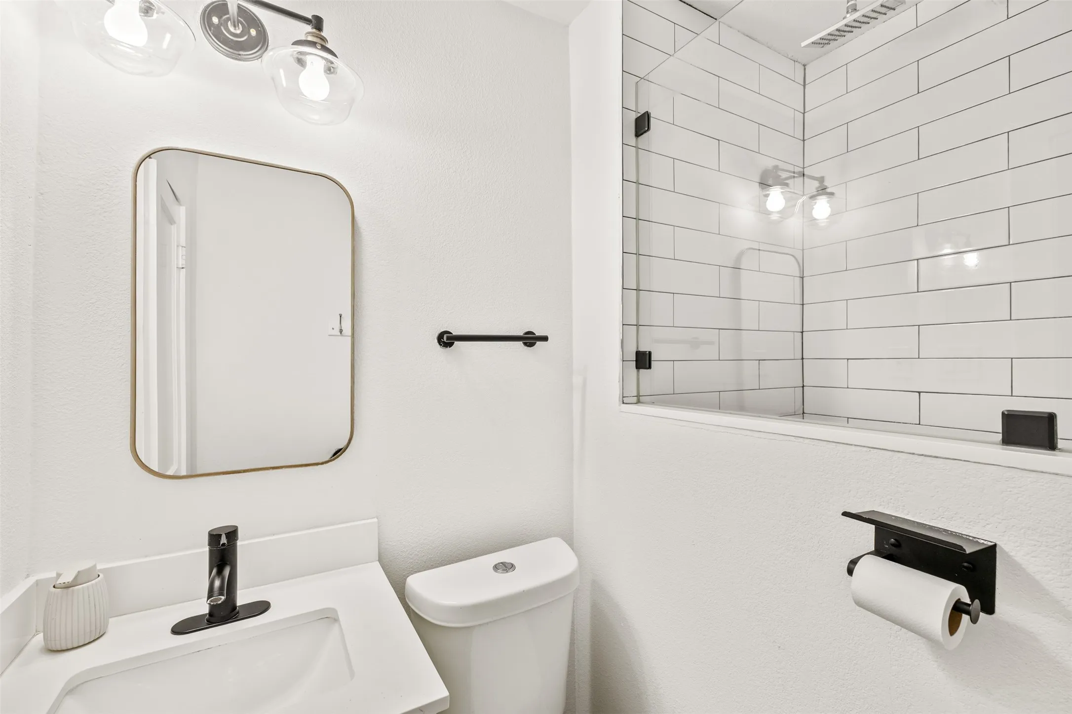 White subway tile shower surround with glass enclosure, white vanity with integrated sink, black single-handle faucet, and black and brass lighting fixtures