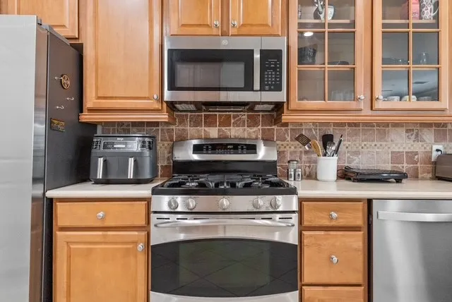 Stainless steel gas range, stainless steel microwave, stone tile backsplash, light wood cabinetry, and stainless steel dishwasher