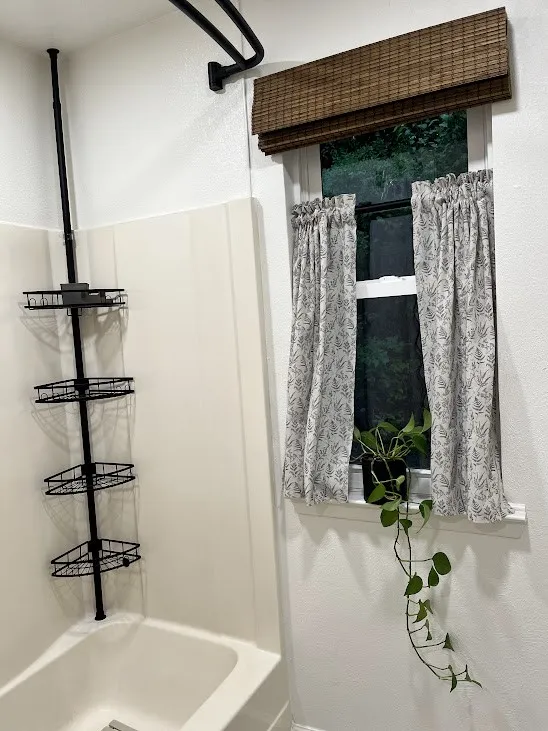 White bathtub with a black shower caddy, white tile surround, and a framed window with a woven wood-finish blind