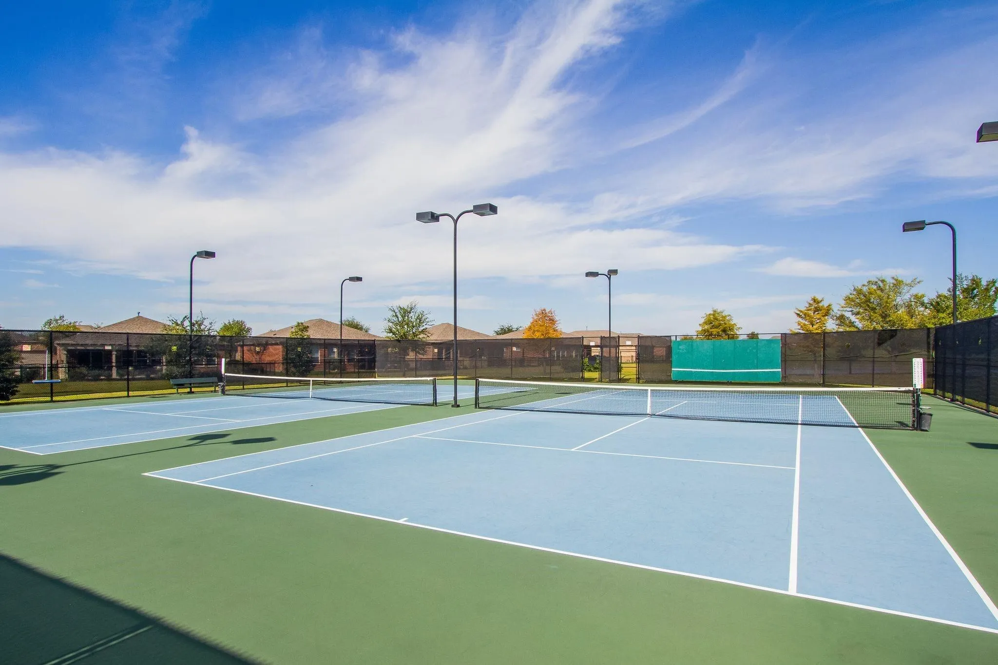 Multiple outdoor tennis courts with light blue playing surfaces and green surrounds, equipped with perimeter fencing and stadium lighting