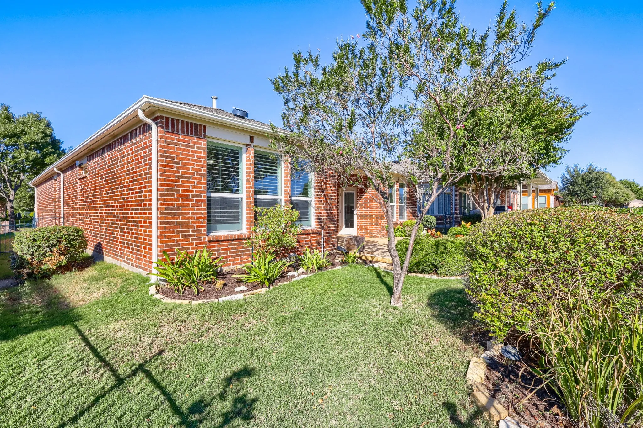 Red brick exterior with white window trim, multiple windows with horizontal blinds, well-maintained lawn, established trees, and perimeter landscaping