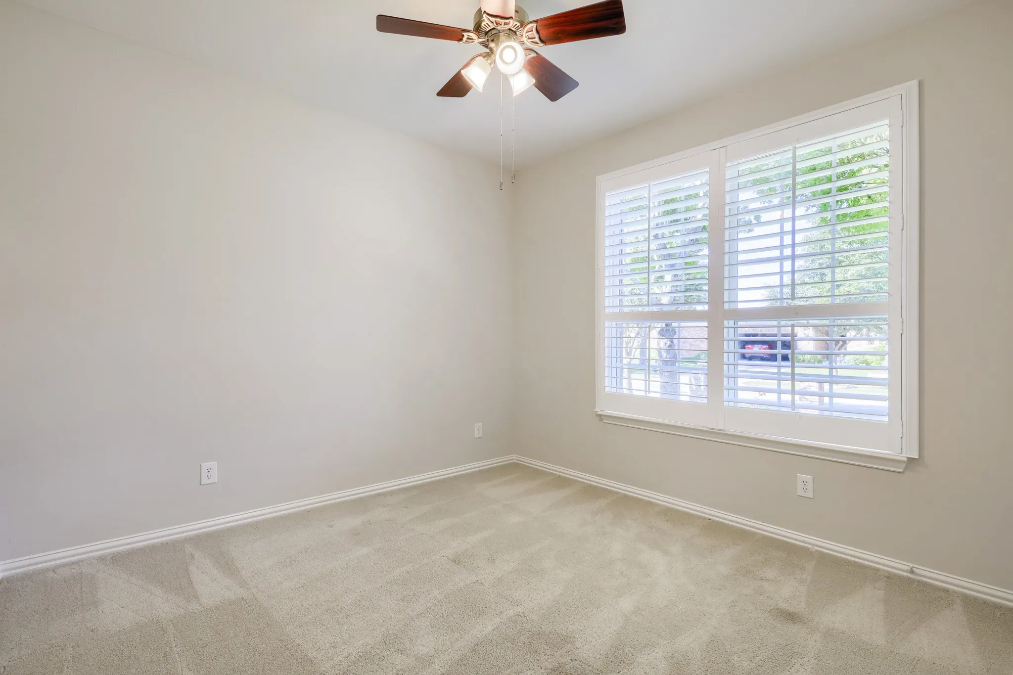 Warm neutral wall color, plush carpet flooring, white plantation shutters, and a ceiling fan with integrated lighting