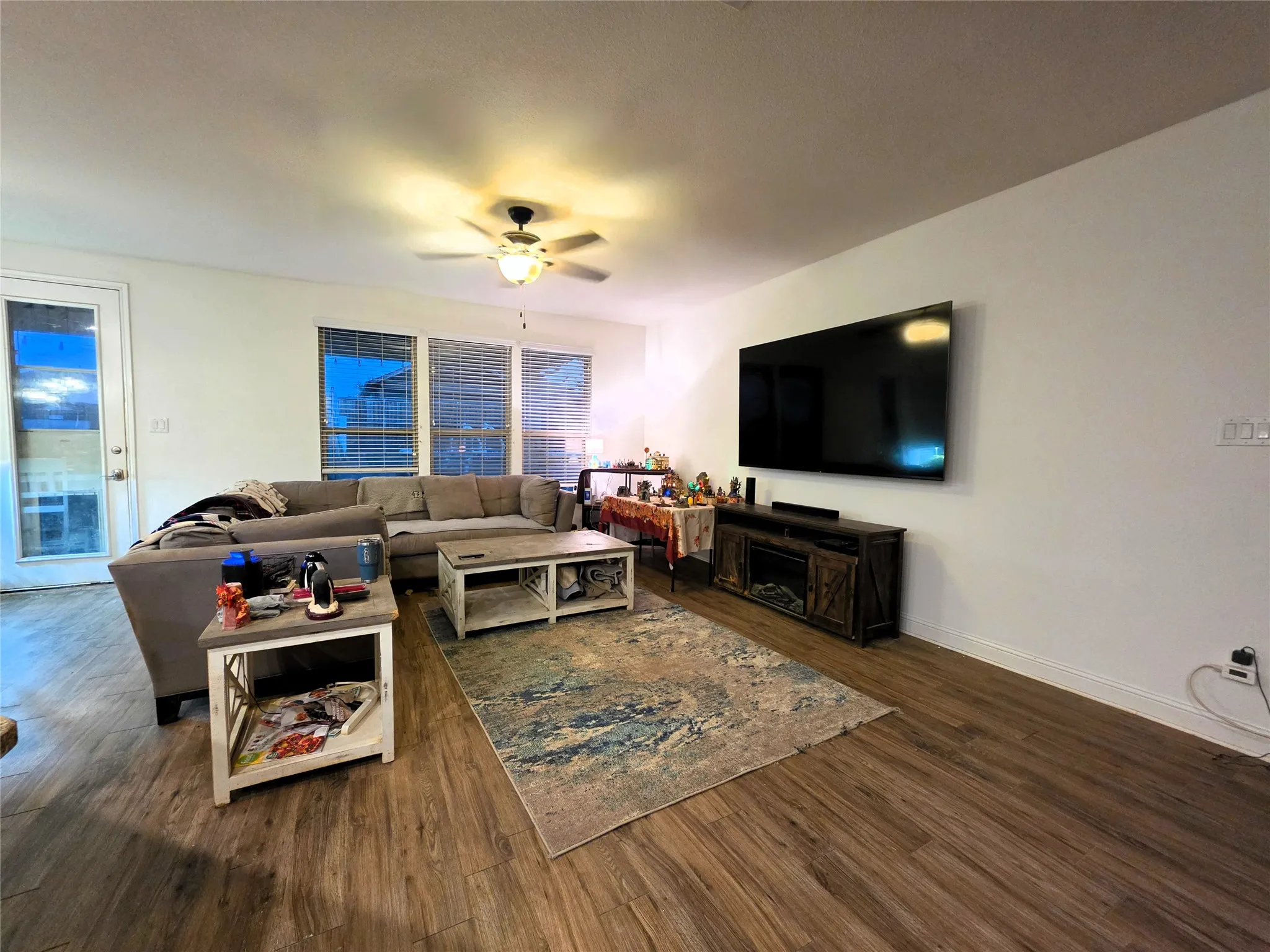 Wood-finish flooring throughout, multiple windows with blinds, a ceiling fan with integrated lighting, and a built-in electric fireplace feature