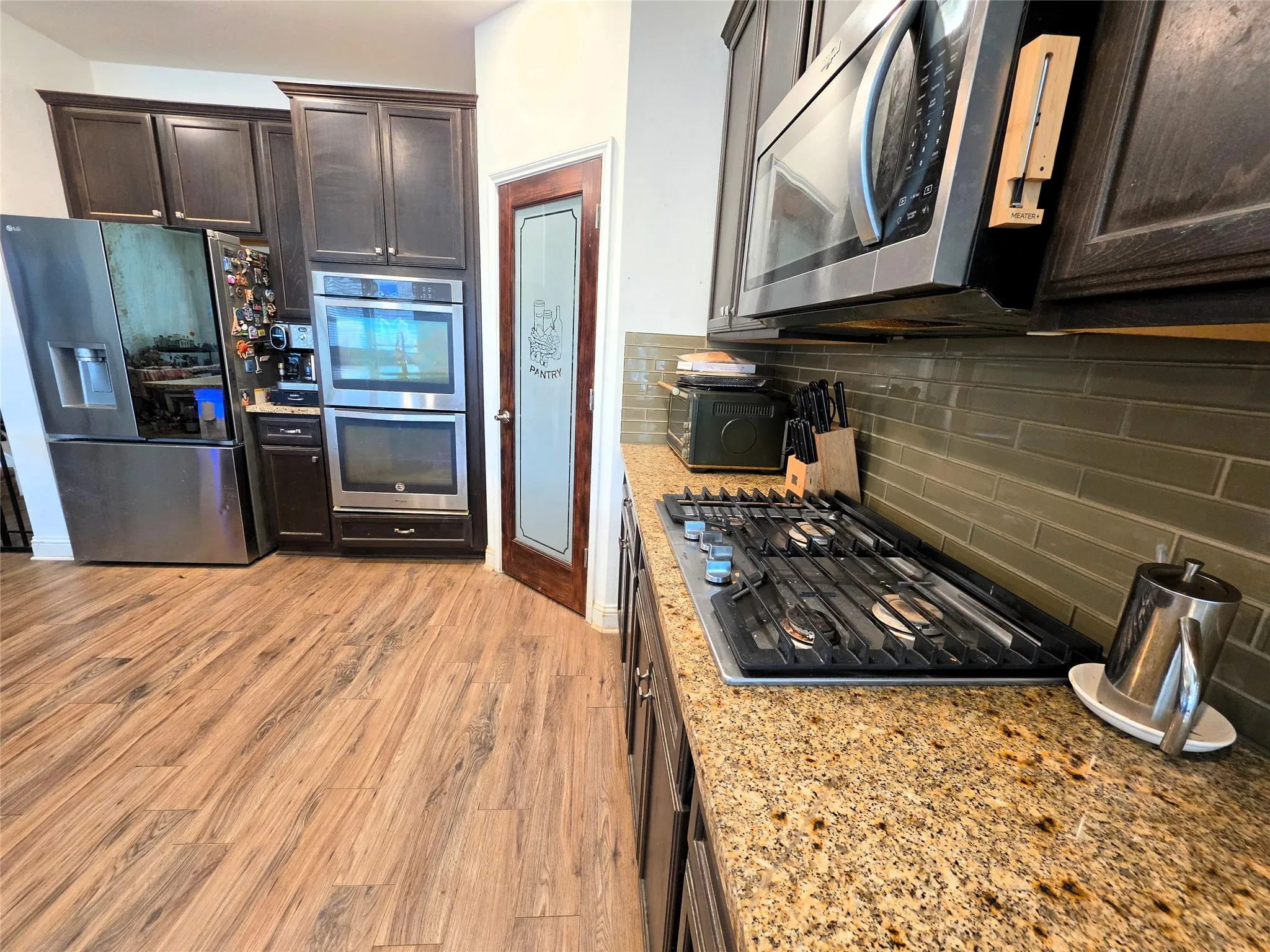 Modern kitchen featuring stainless steel appliances including a gas cooktop and double wall ovens, granite countertops, dark cabinetry with hardware accents, subway tile backsplash, and wood-finish flooring throughout the open layout