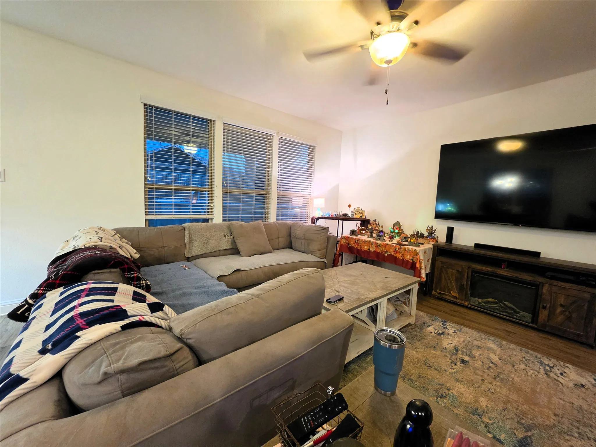 Wood-finish flooring, multiple windows with blinds, ceiling fan with integrated lighting, and a built-in media console with a fireplace insert