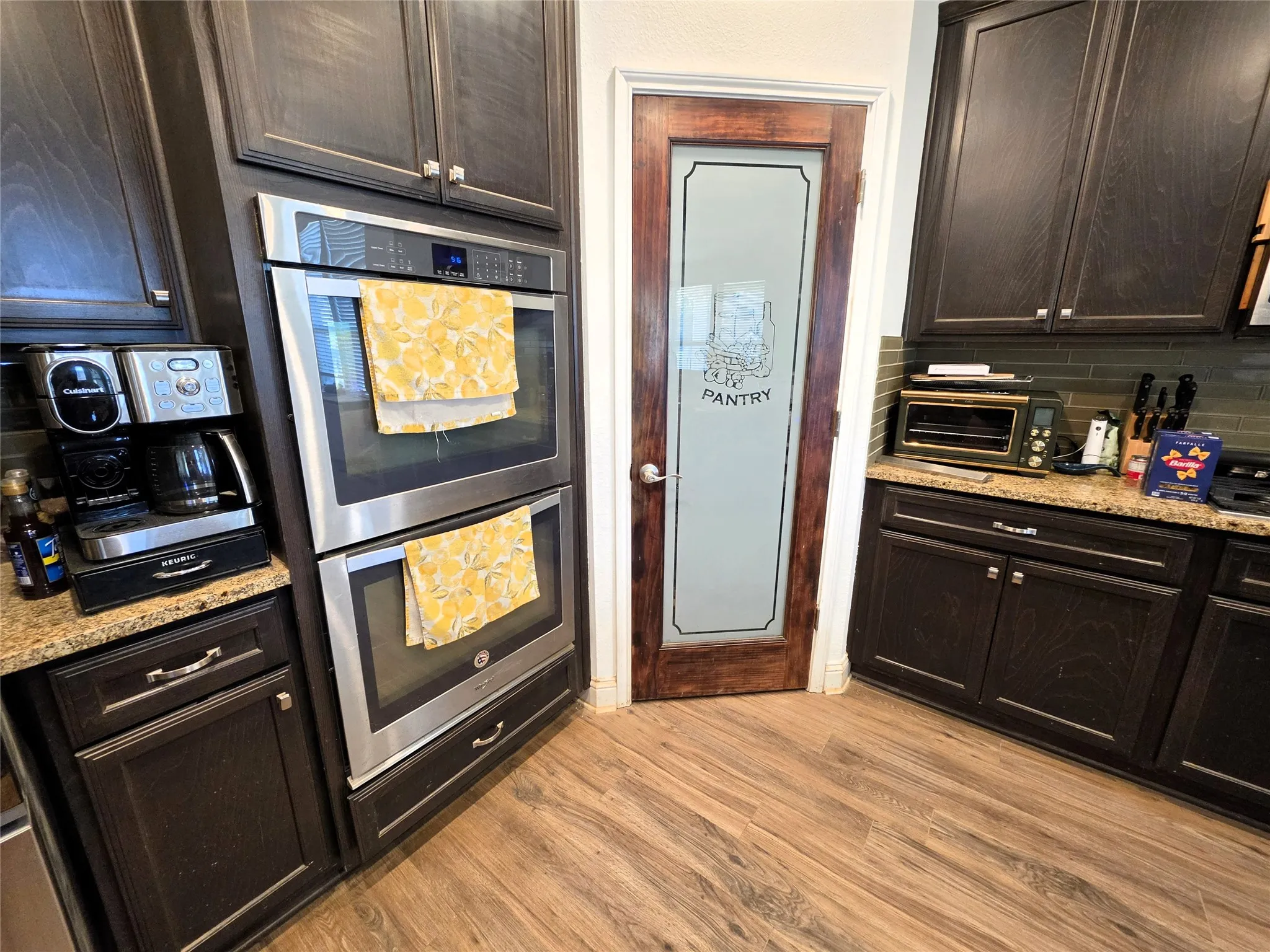 Wood-finish flooring, dark wood cabinetry with silver-tone hardware, granite-style countertops, stainless steel double wall ovens, and a pantry door with frosted glass panel