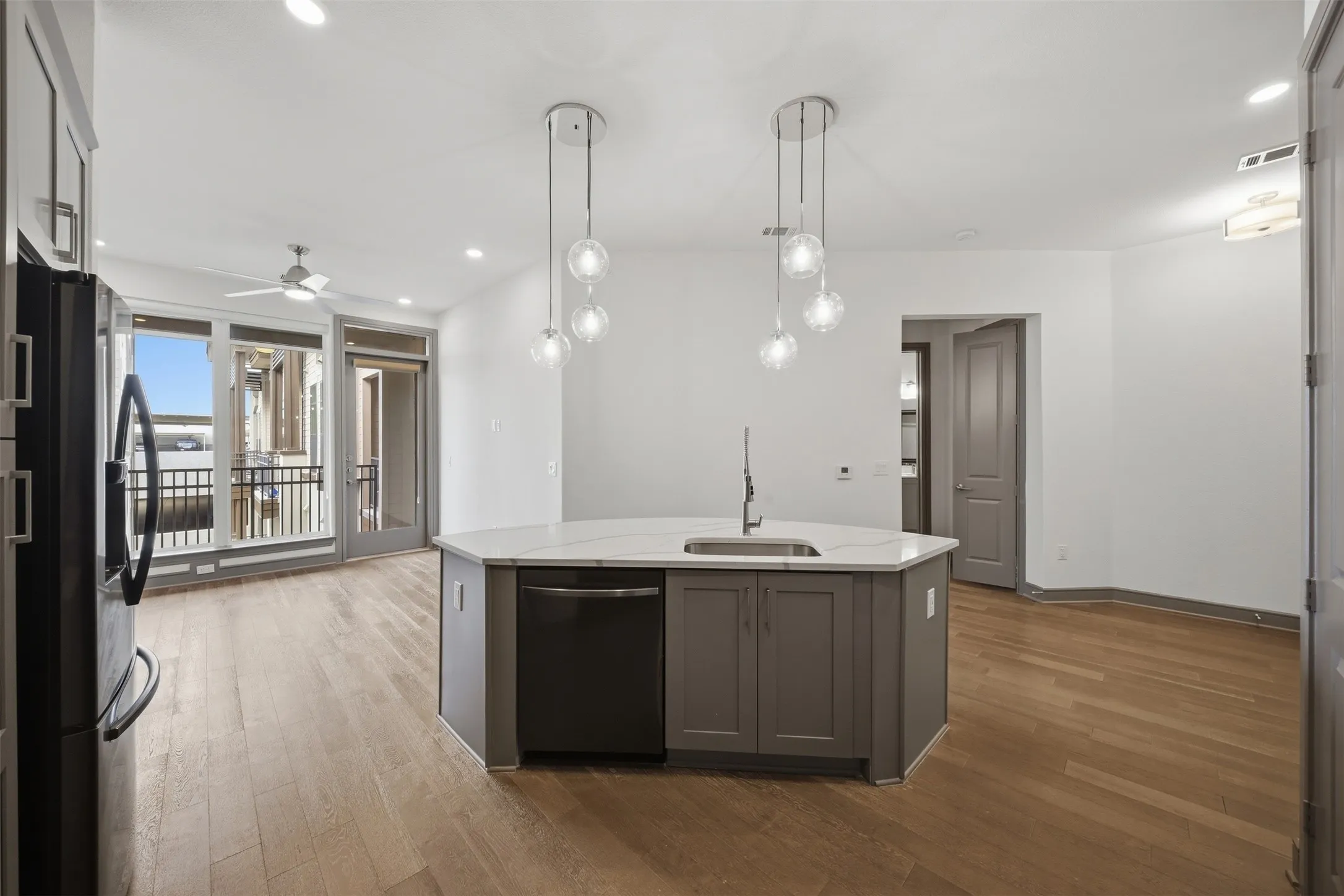Open-concept living area featuring wood-finish flooring, a kitchen island with integrated sink, two multi-bulb pendant lights, and large glass-paned doors opening to a balcony