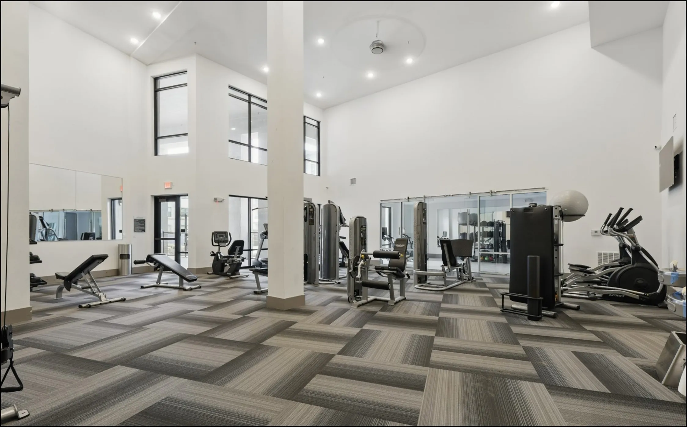 Spacious fitness center featuring varied exercise equipment, a mirrored wall, high ceilings, large windows, and patterned carpet flooring