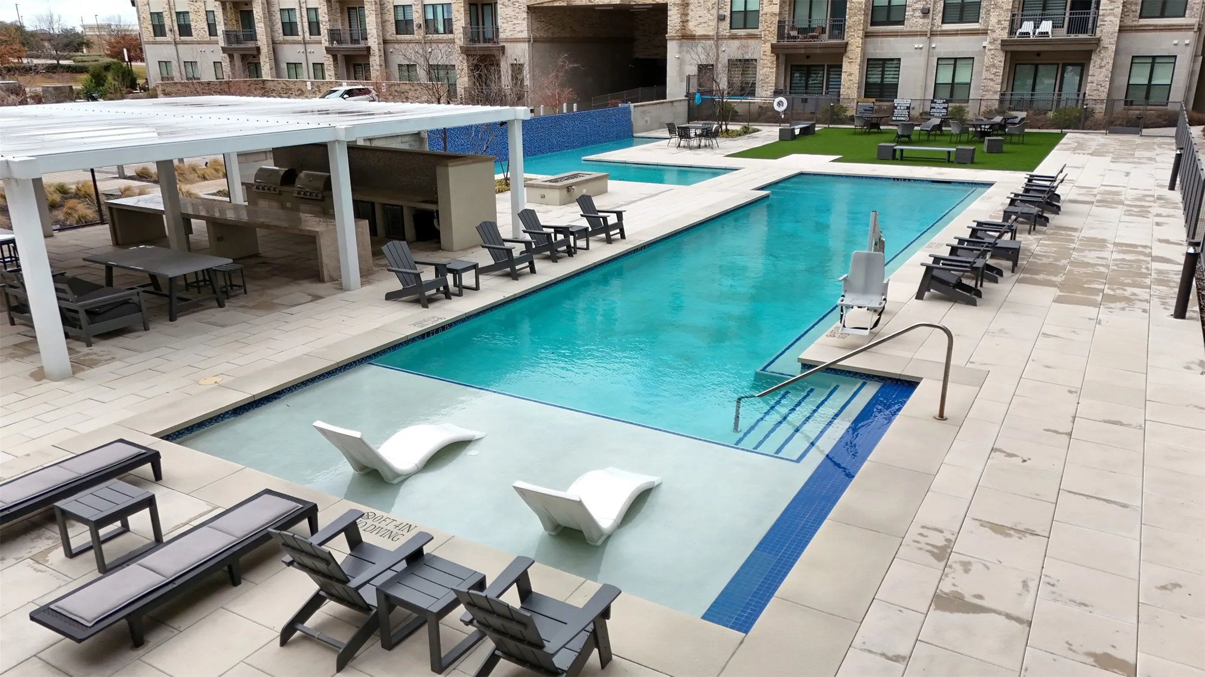Expansive pool area featuring a perimeter fence, a large swimming pool with integrated sun shelves, an outdoor kitchen with multiple grills, a shaded pergola, and a separate turf lounge area