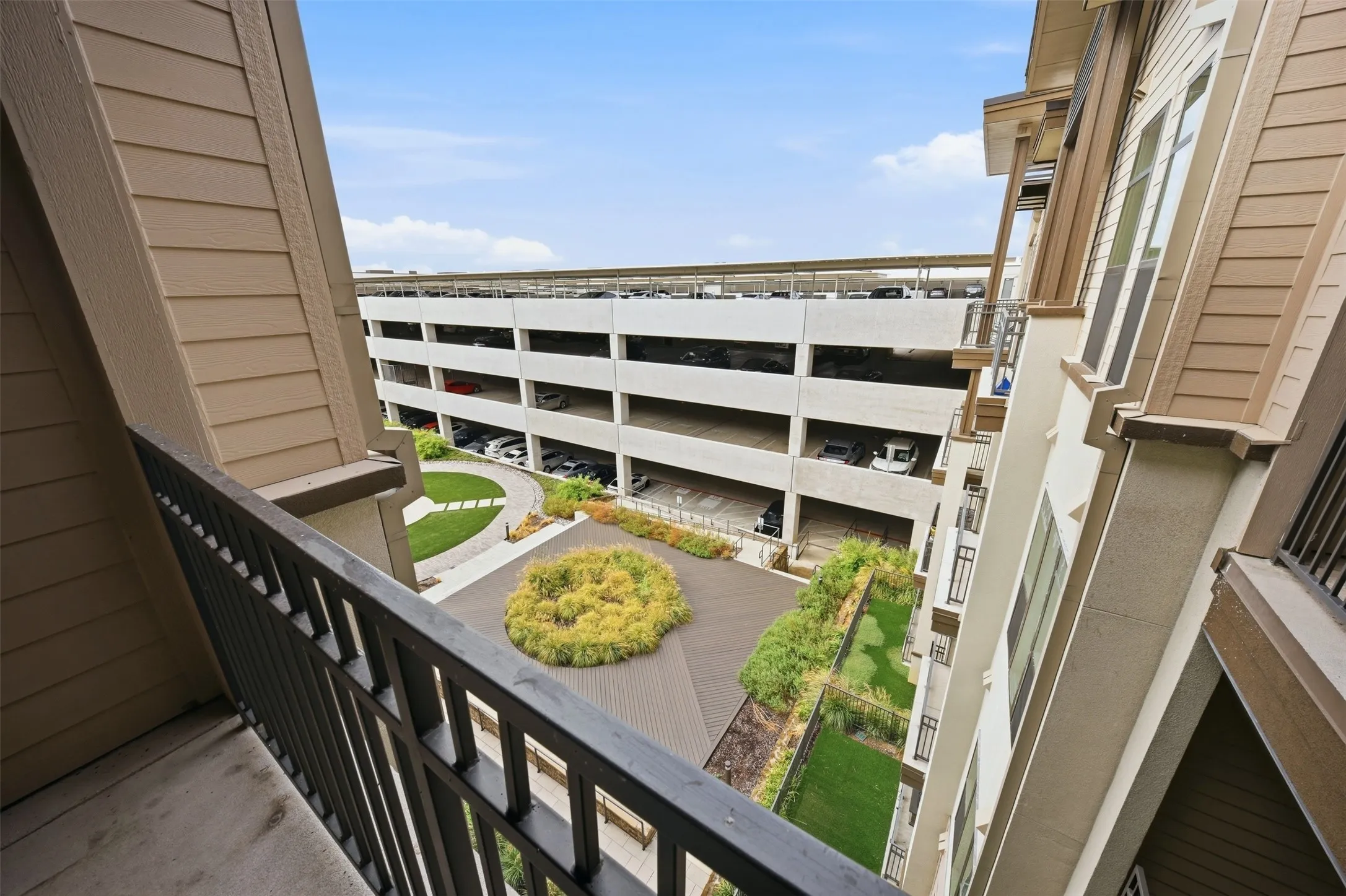 Private balcony with metal railing, offering views of the multi-level parking structure and a landscaped courtyard featuring a circular lawn and ground cover plantings