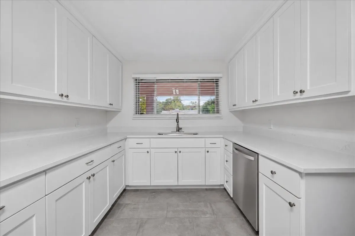 White shaker-style cabinetry, light-toned countertops, stainless steel dishwasher, and a single basin sink with a chrome faucet
