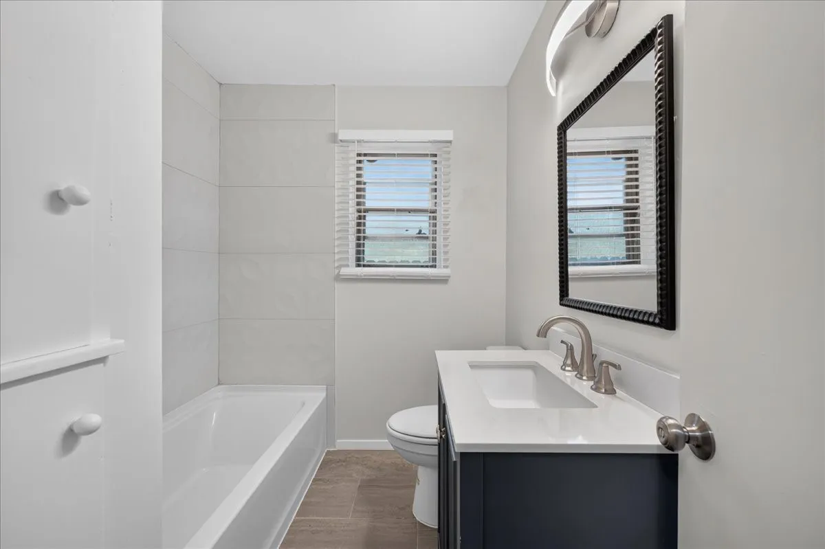 Bathtub with white subway tile surround, dark wood-finish vanity with a white countertop and integrated sink, brushed nickel faucet, and a black framed mirror