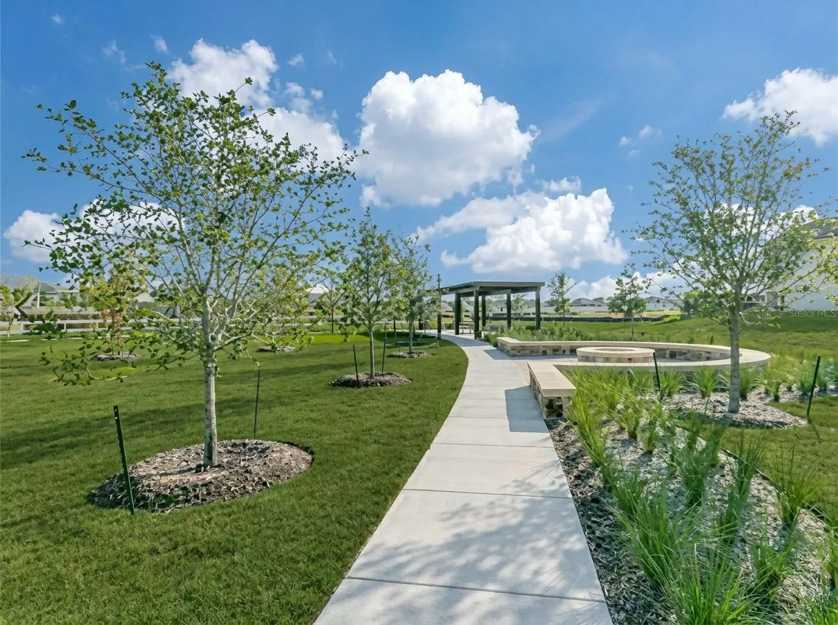 Manicured lawn with a concrete pathway leading to a stone-clad fire pit, surrounded by lush landscaping, and a dark-frame pergola