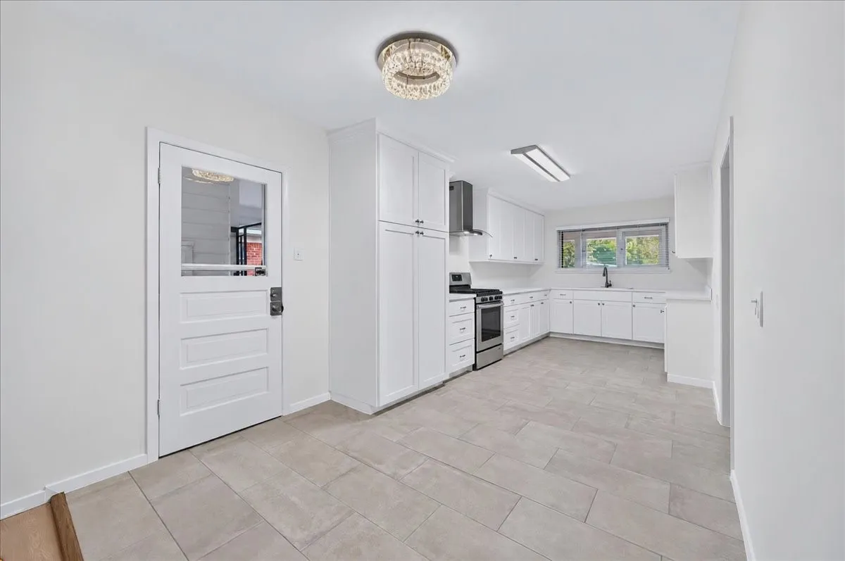 Open kitchen featuring white cabinetry, light-toned tile flooring, stainless steel appliances, a built-in range hood, and a window above the sink