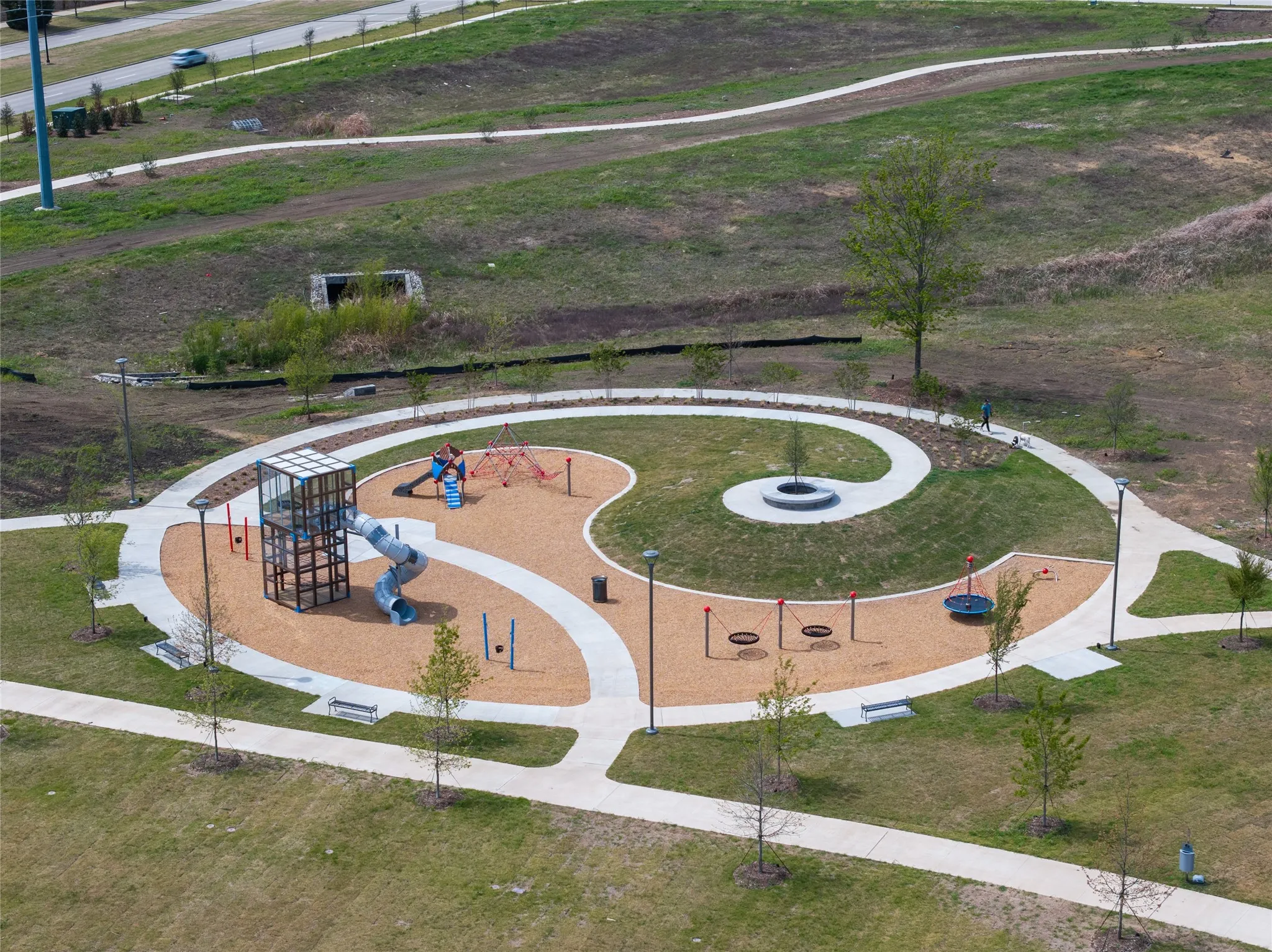 Recreational area featuring a spiral playground structure, sand-colored ground cover, paved walkways, and integrated park benches