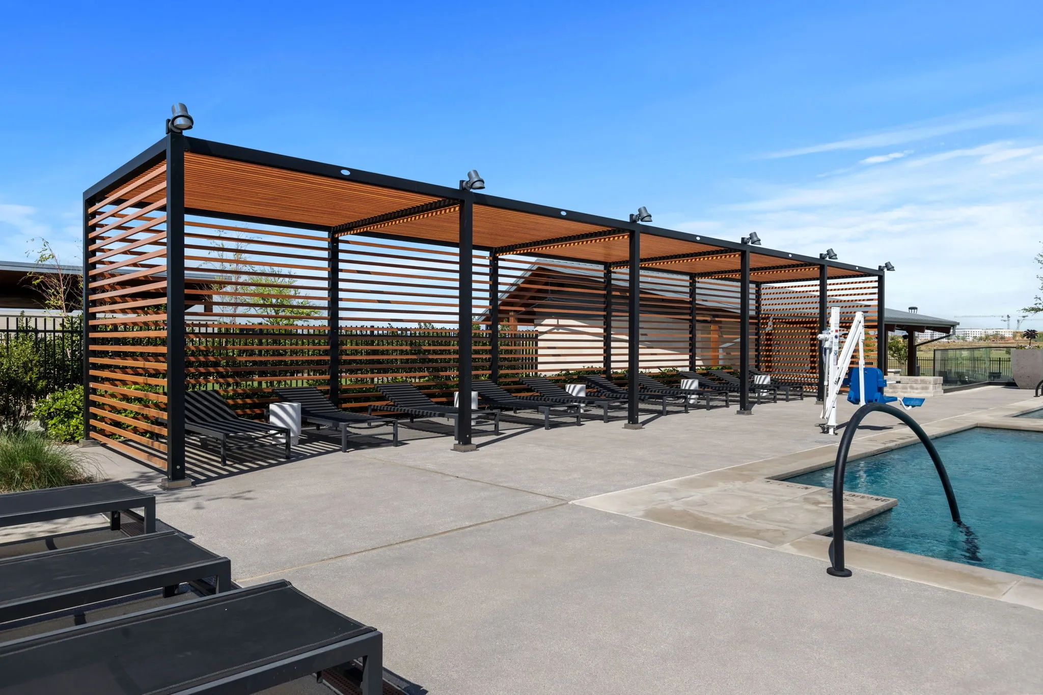 Expansive poolside concrete patio featuring a multi-section pergola with wood-finish slatted sides and roof, and a swimming pool with a dark-finished handrail