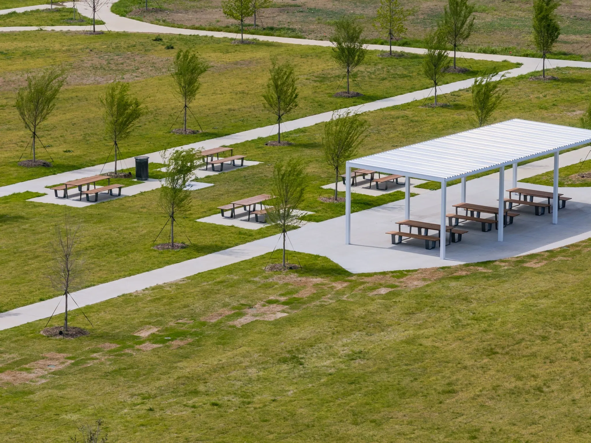 Expansive green space featuring a white metal pergola, multiple picnic tables, paved walking paths, and young trees