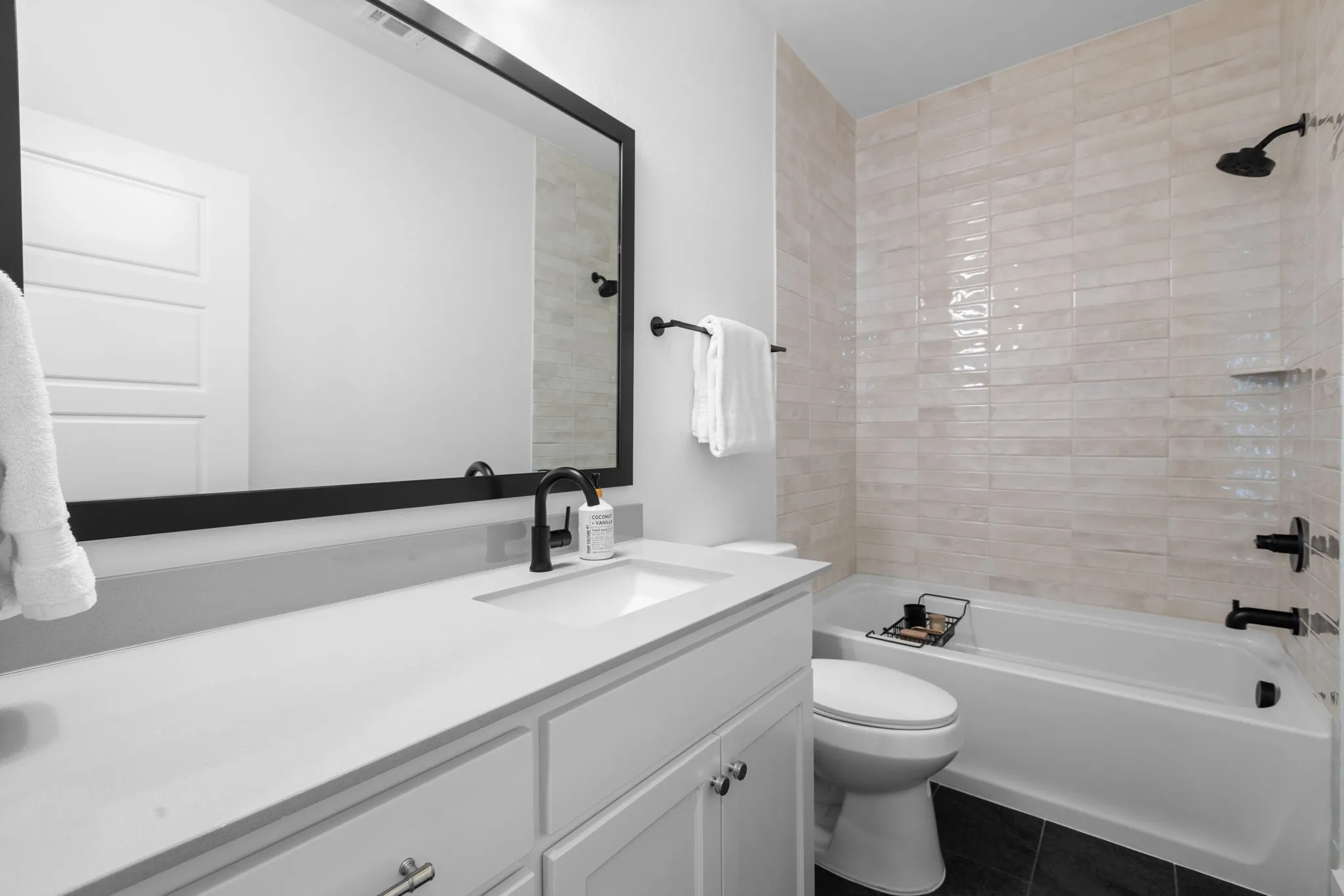 White vanity with integrated sink, black framed mirror, matte black fixtures, full tile tub surround, and black tile flooring
