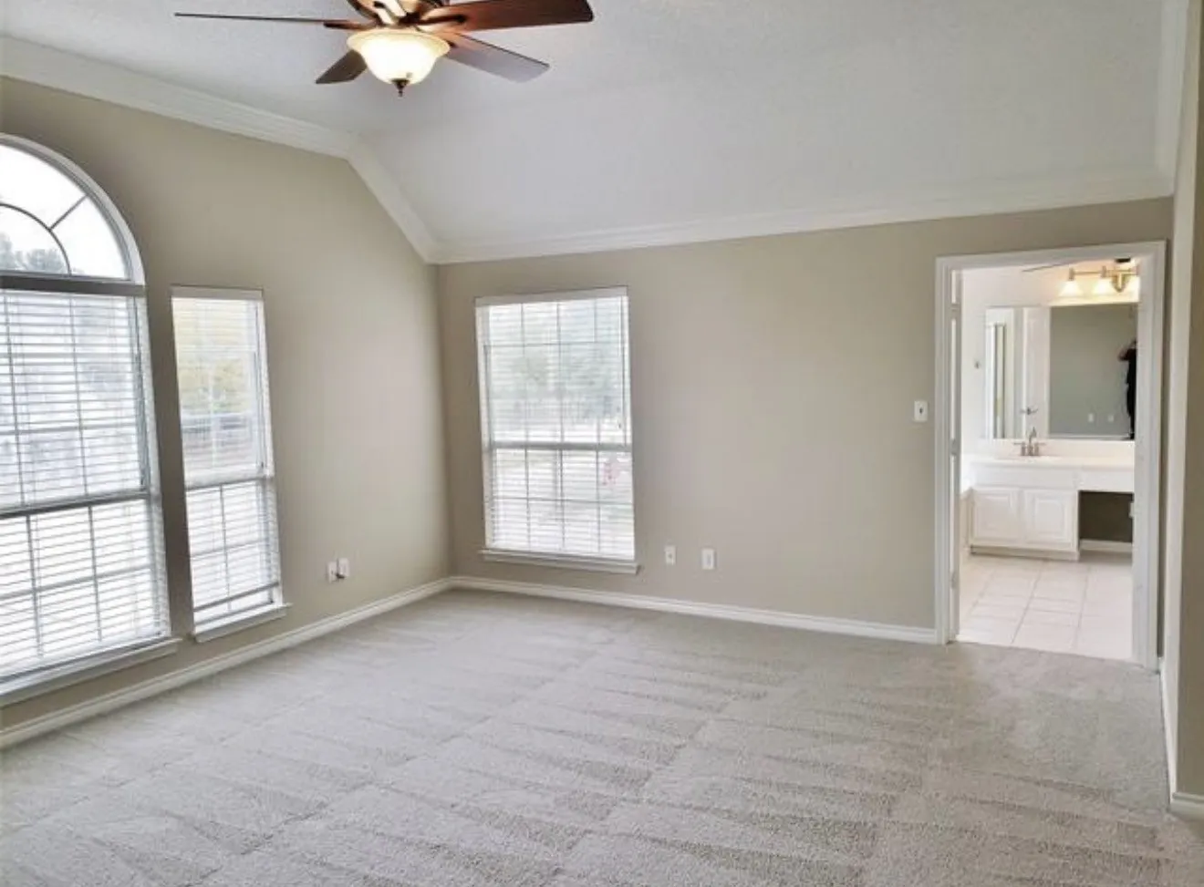 Vaulted ceiling with crown molding, ceiling fan, arched transom window, carpeted flooring, and an adjoining bathroom with a vanity