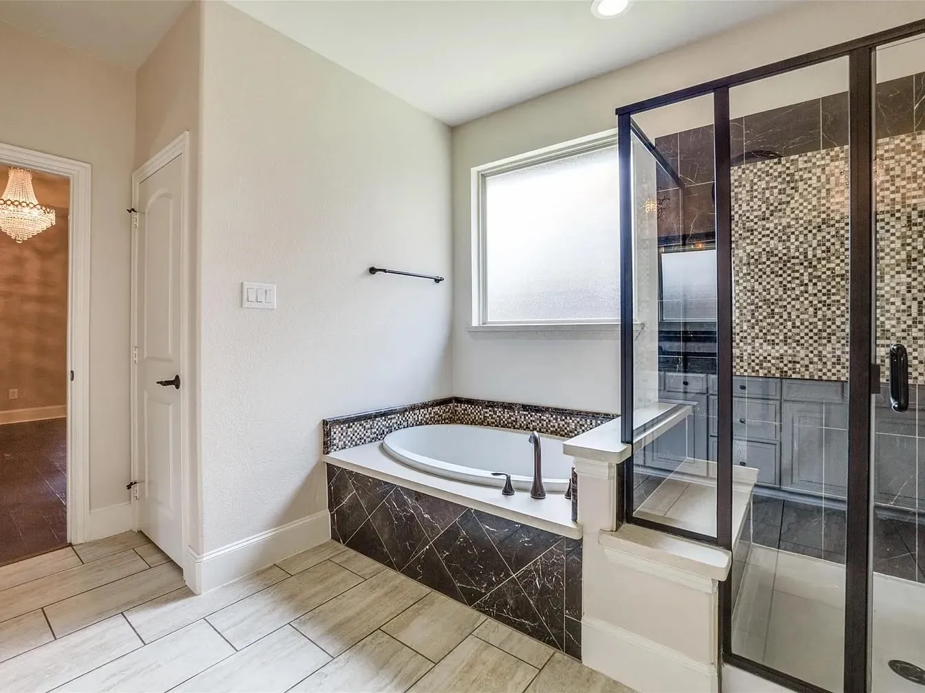 Built-in soaking tub with dark marble-finish surround, glass-enclosed shower with mosaic tile accent, and light-toned tile flooring
