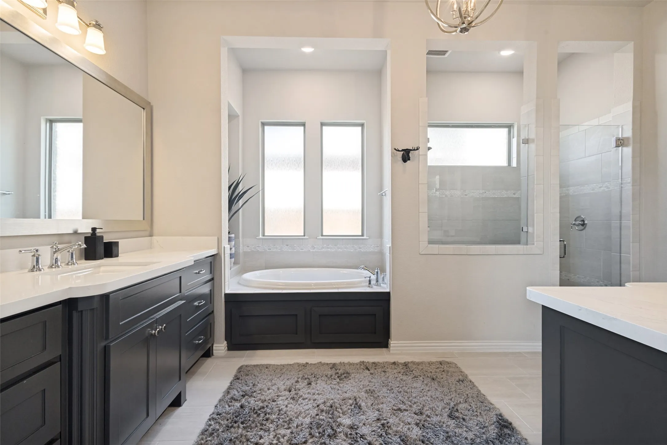 Spacious primary bathroom featuring twin vanities with dark cabinetry and light countertops, a built-in soaking tub, and a glass-enclosed shower with tiled walls.
