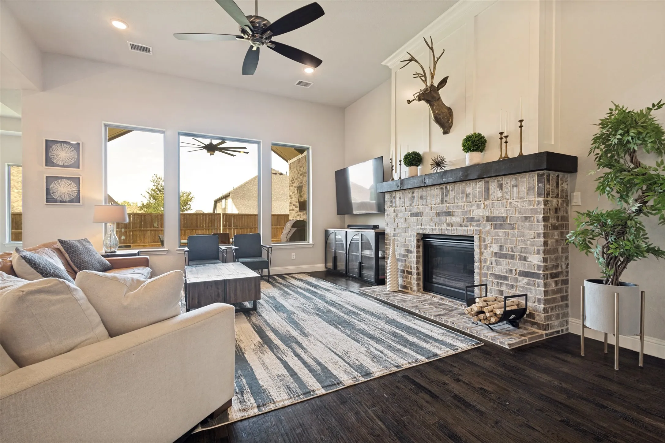 Spacious living area featuring a brick fireplace with a dark wood mantel, high ceilings with a ceiling fan, dark wood-finish flooring, and multiple large picture window with views of the large backyard patio and private backyard