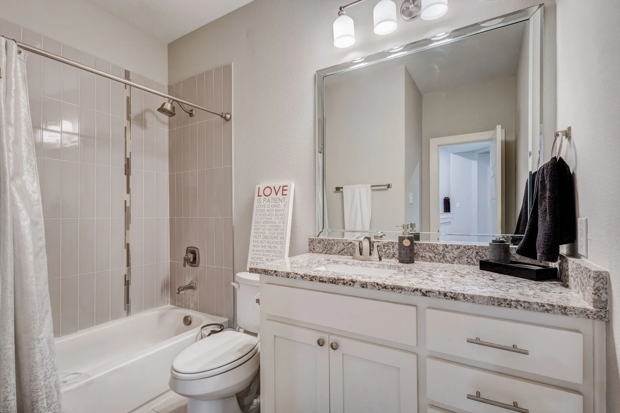 Bathtub with vertical tile surround, vanity with granite countertop, undermount sink, white cabinetry with silver hardware, and a large wall-mounted mirror