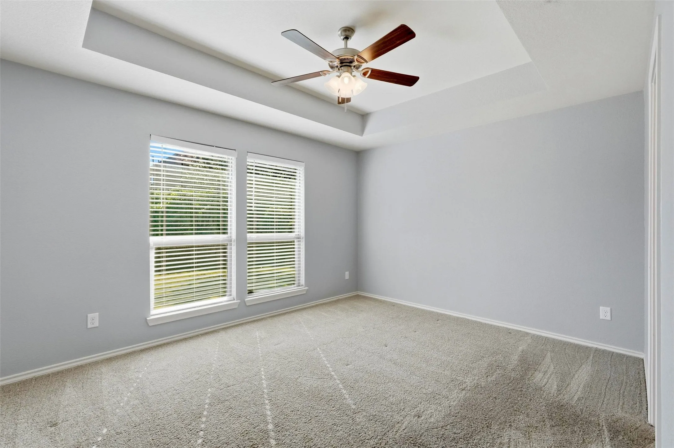 Tray ceiling with a ceiling fan light fixture, dual windows with blinds, and soft gray carpeting