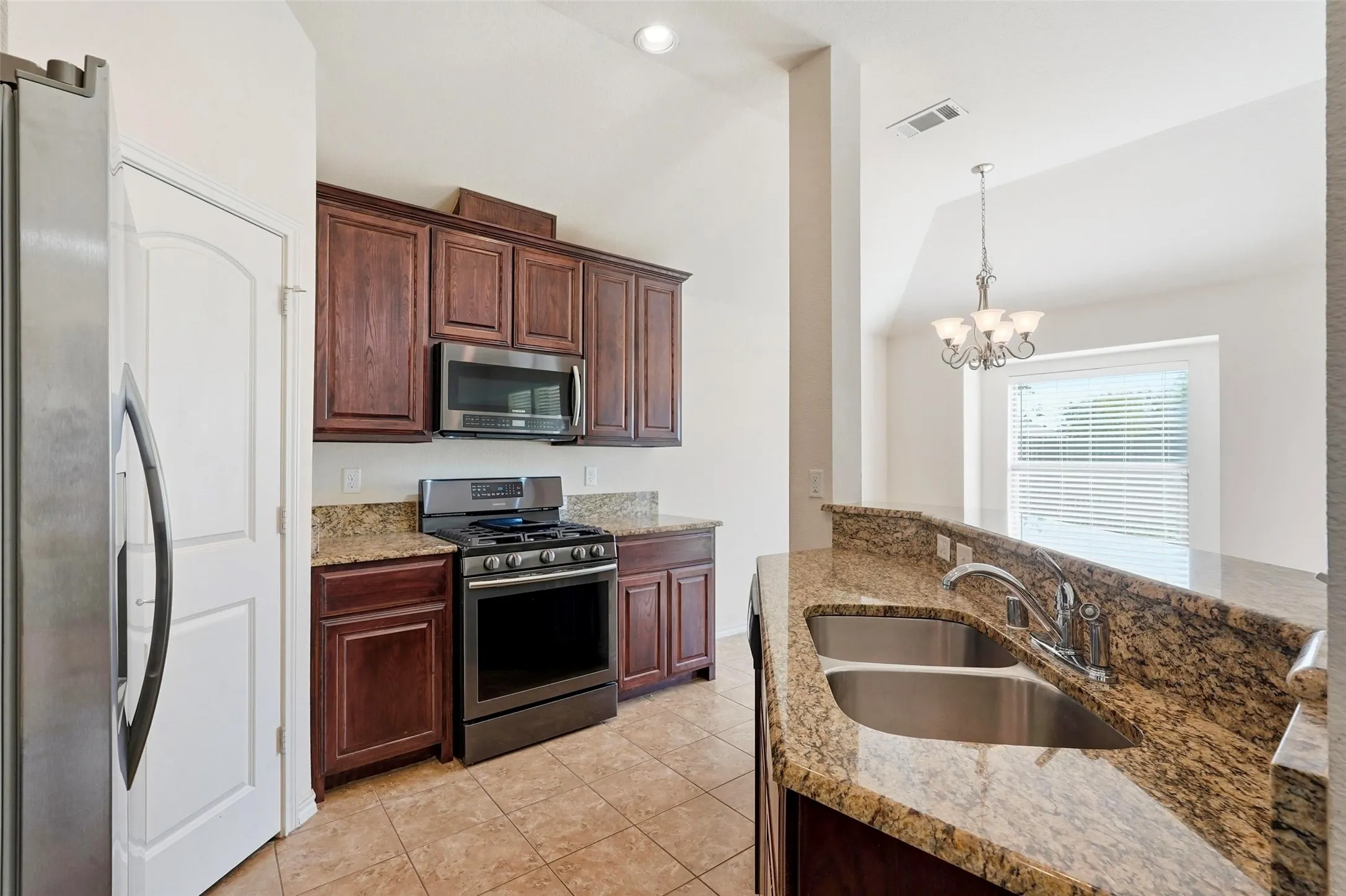 Kitchen featuring rich wood cabinetry, stainless steel appliances, speckled stone countertops, a double basin sink, and tile flooring