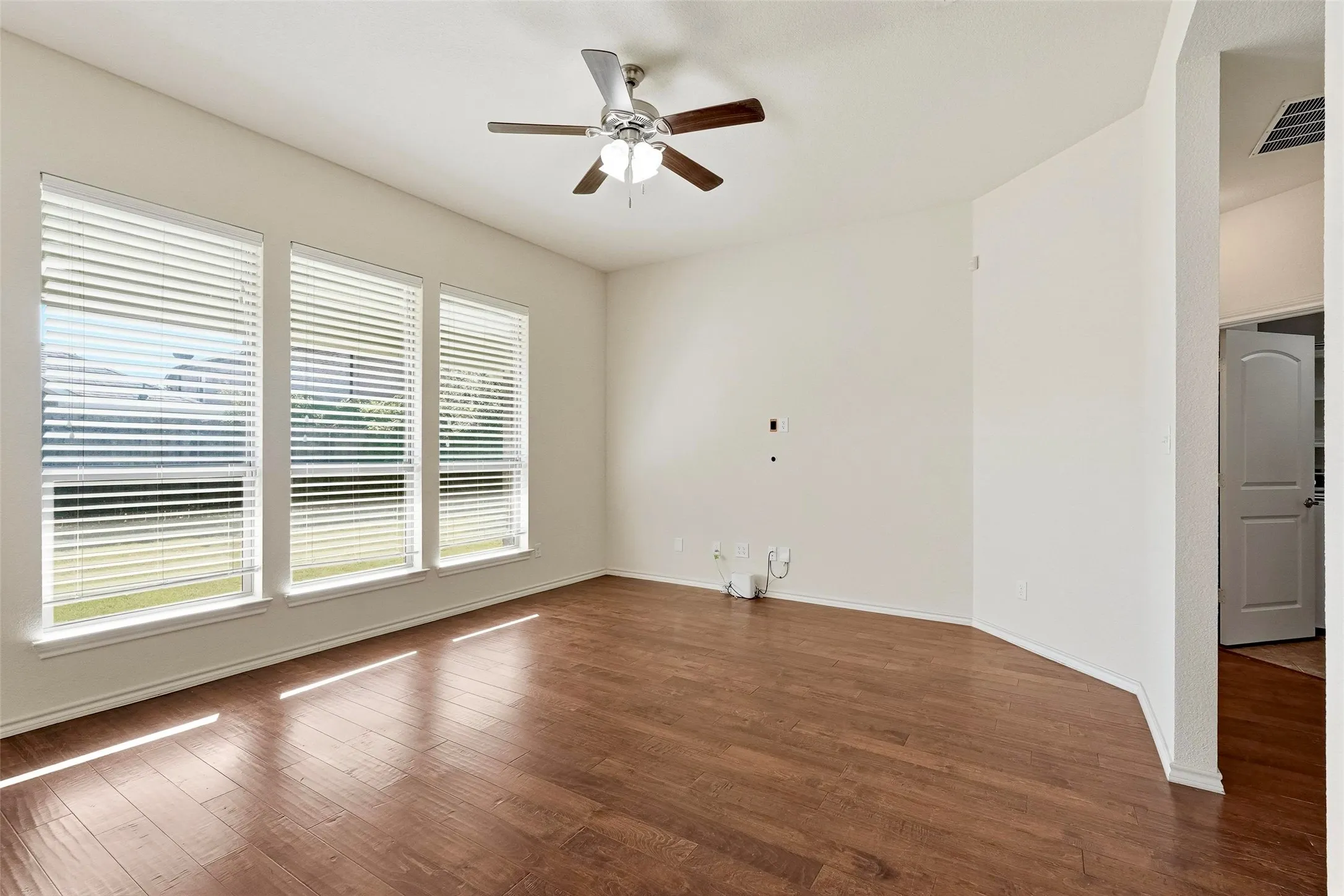 Wood-finish flooring, three large windows with blinds, ceiling fan, and white walls