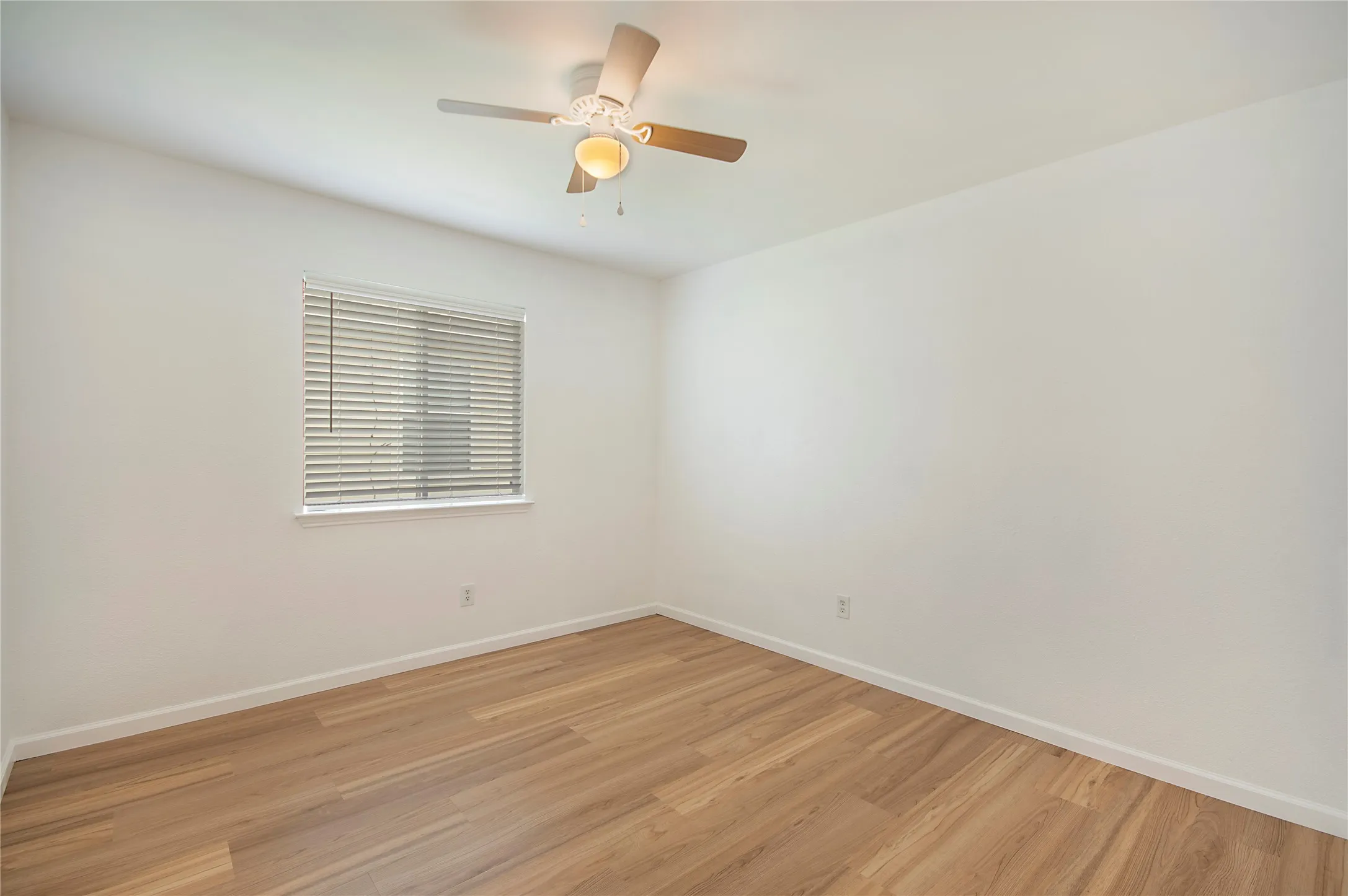 Light wood-finish flooring, white walls, window with blinds, and ceiling fan with integrated lighting