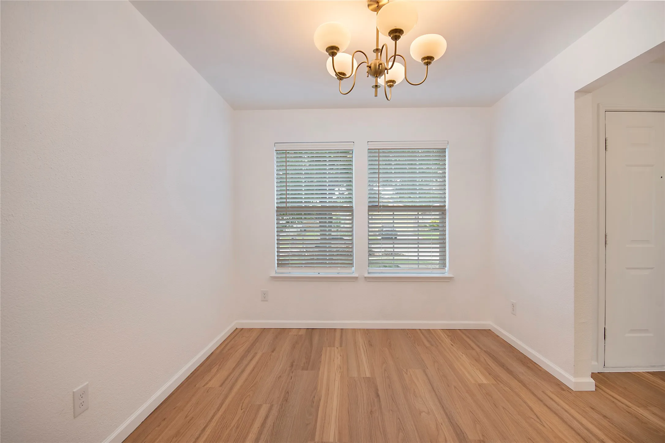 Wood-finish flooring, dual windows with blinds, white painted walls, and a brass-tone chandelier with frosted glass shades