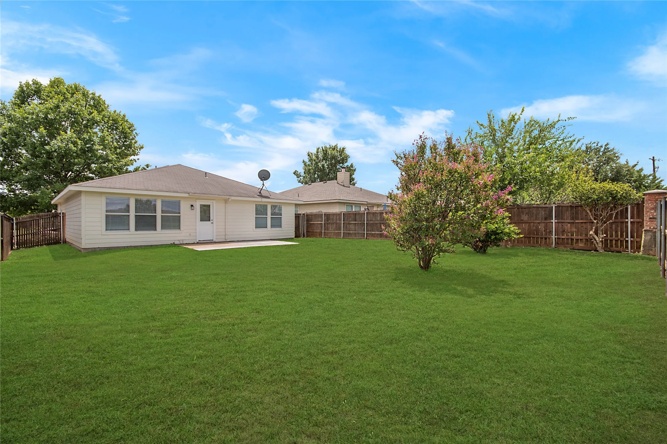 Expansive grassy yard featuring a wood privacy fence, concrete patio, and a mature tree with pink blooms