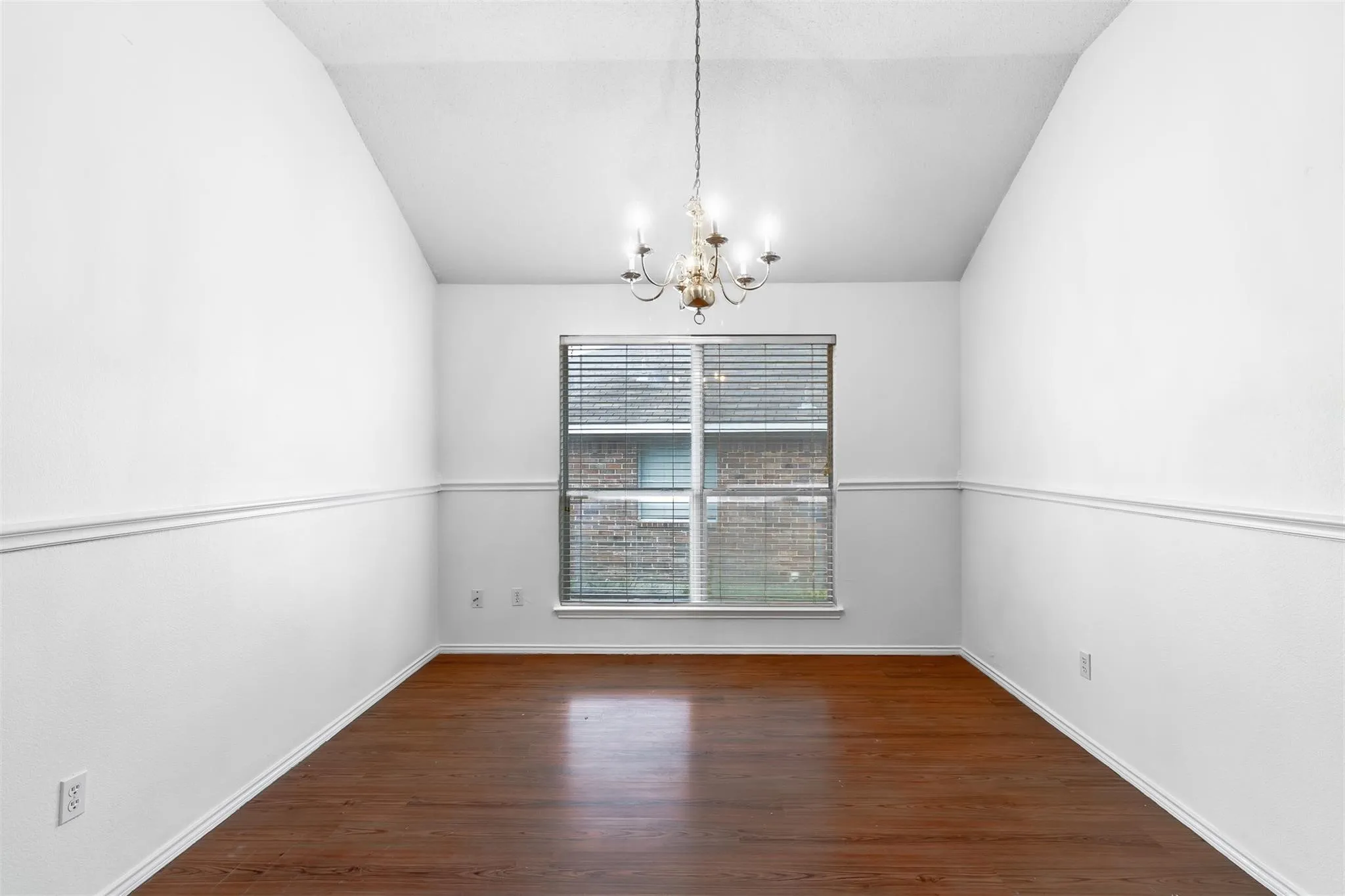 Vaulted ceiling with an ornamental chandelier, rich wood-finish flooring, and a window with horizontal blinds
