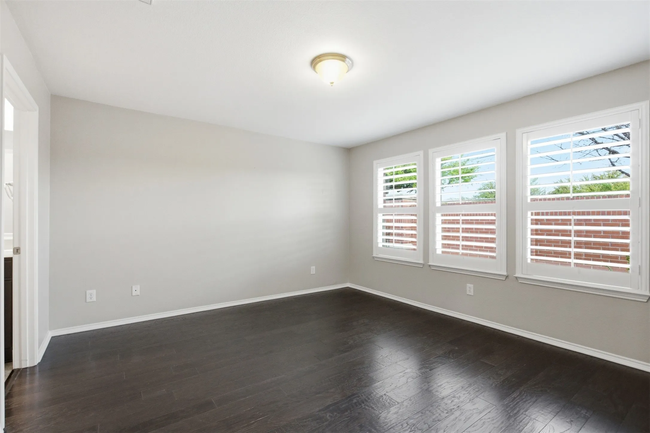 Three windows with white plantation shutters, dark wood-finish flooring, light-toned wall paint, and a semi-flush mount ceiling light