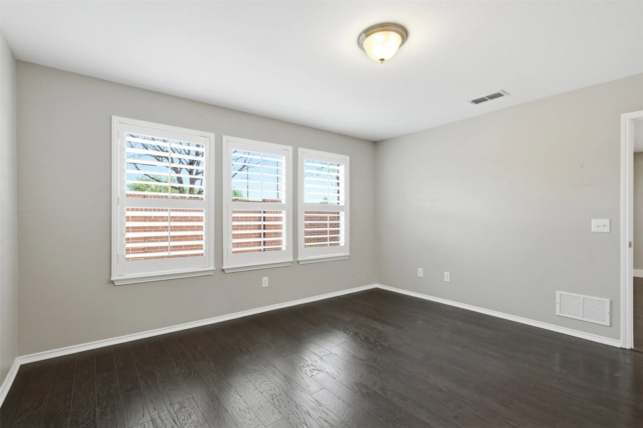 Dark wood-finish flooring, three windows with white plantation shutters, and a ceiling-mounted light fixture