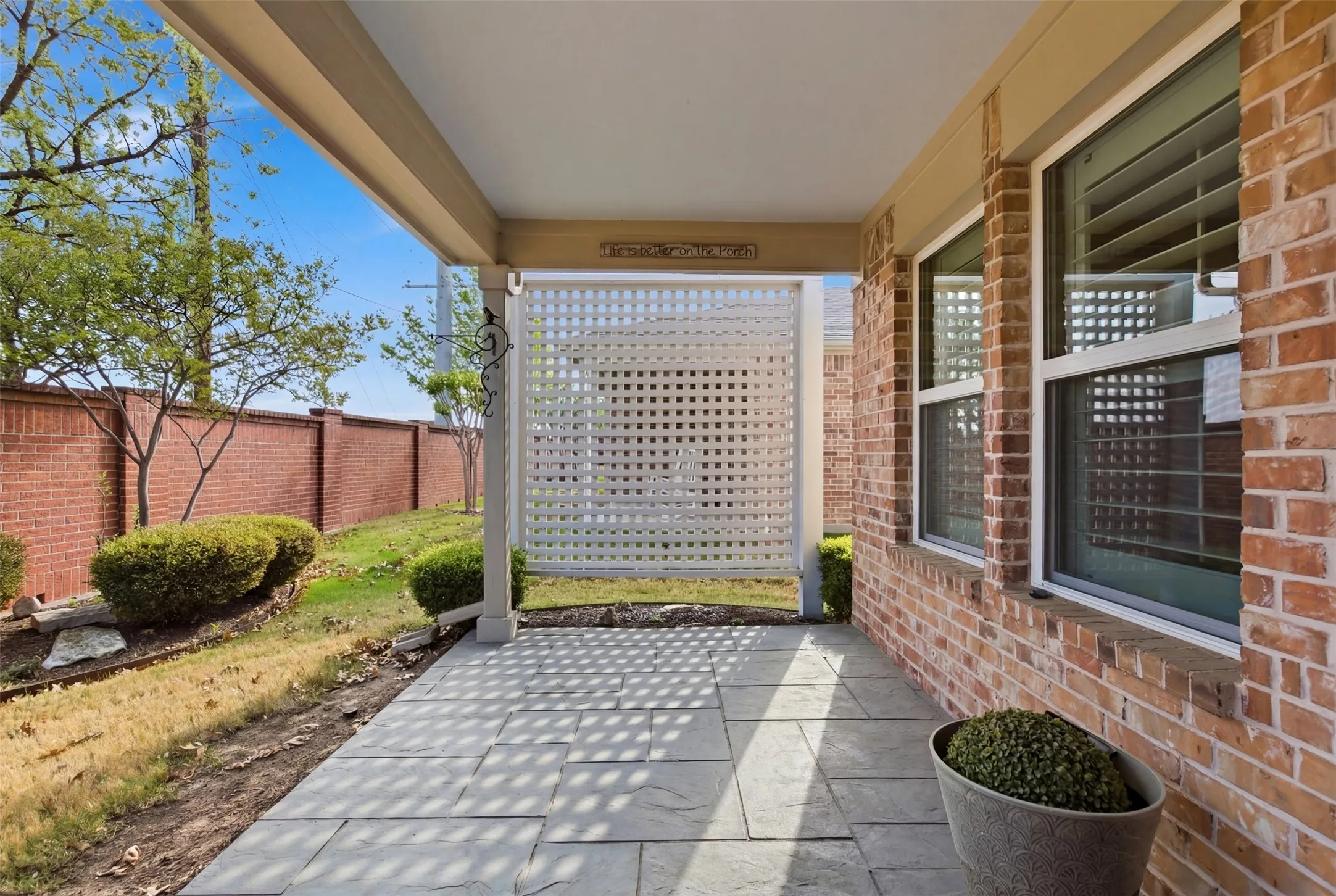 Covered patio with stone tile flooring, brick exterior, white lattice privacy screen, and multiple windows