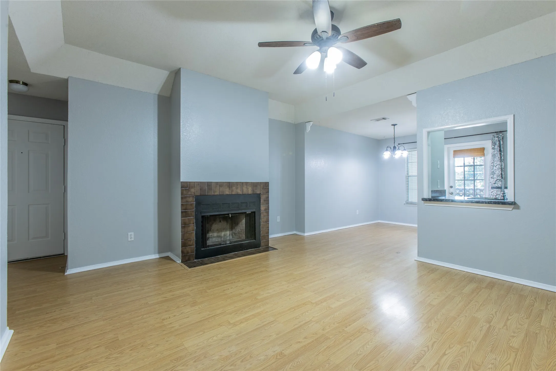 Living area featuring wood-finish flooring, a ceiling fan with integrated lighting, a fireplace with a tile surround, and a pass-through window with a countertop