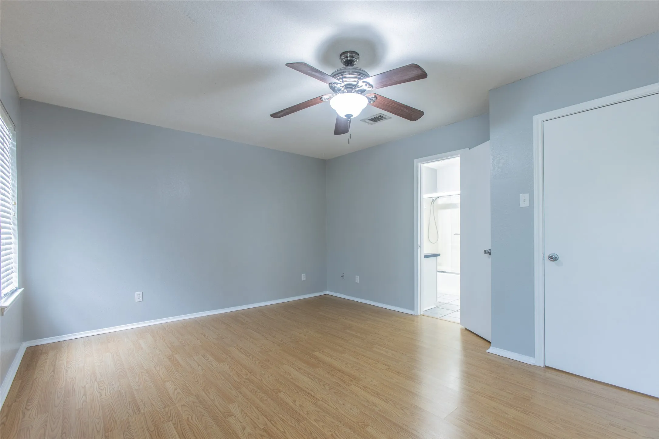 Wood-finish flooring, ceiling fan with integrated lighting, window with horizontal blinds, white interior doors, and pale blue wall finish