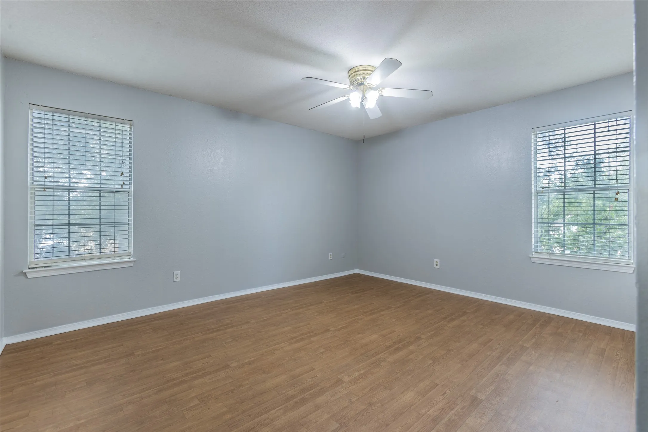 Spacious interior with wood-finish flooring, two windows with blinds, a ceiling fan, and light gray walls