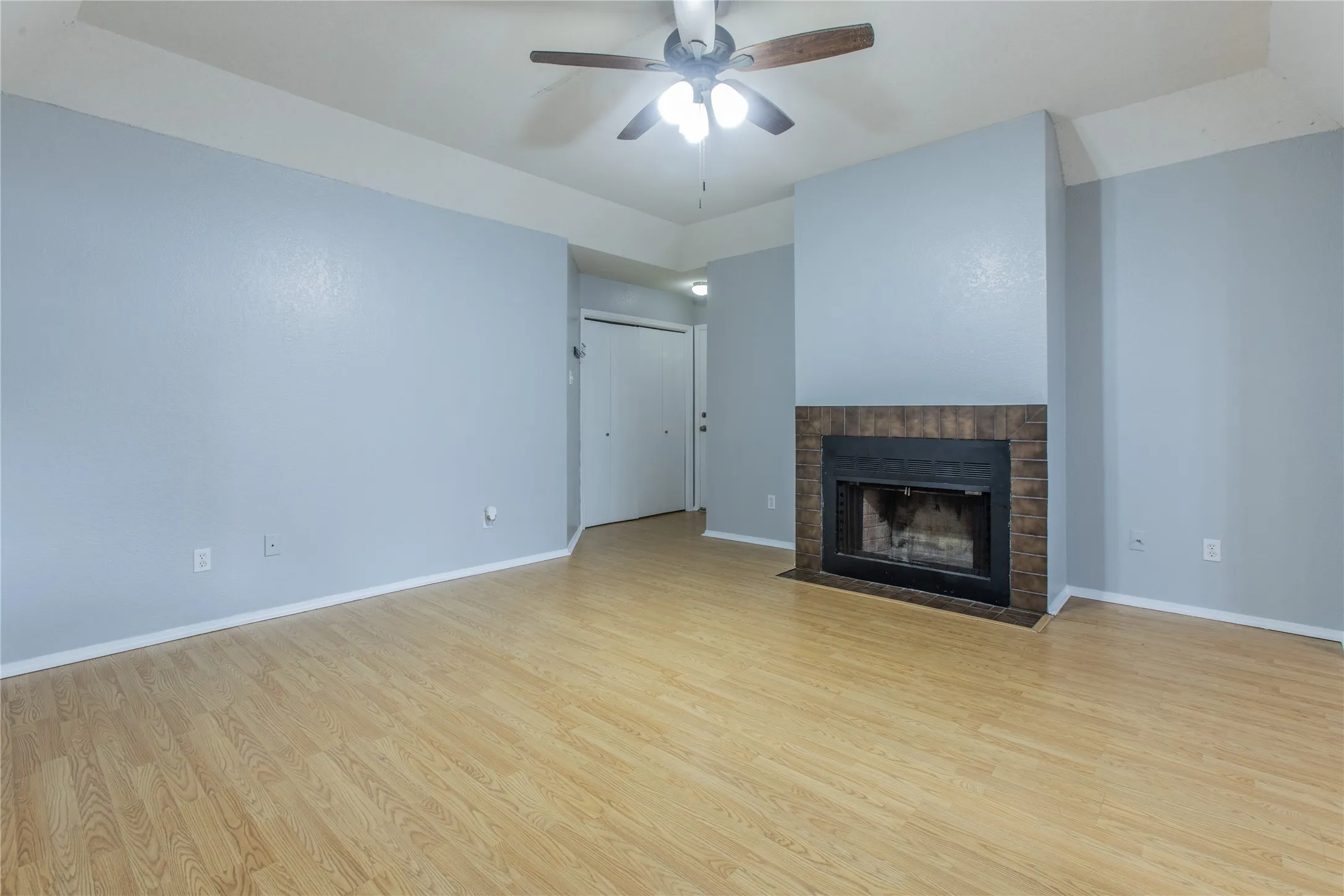 Wood-finish flooring, brick-surround fireplace, ceiling fan with integrated lighting, and bi-fold closet doors