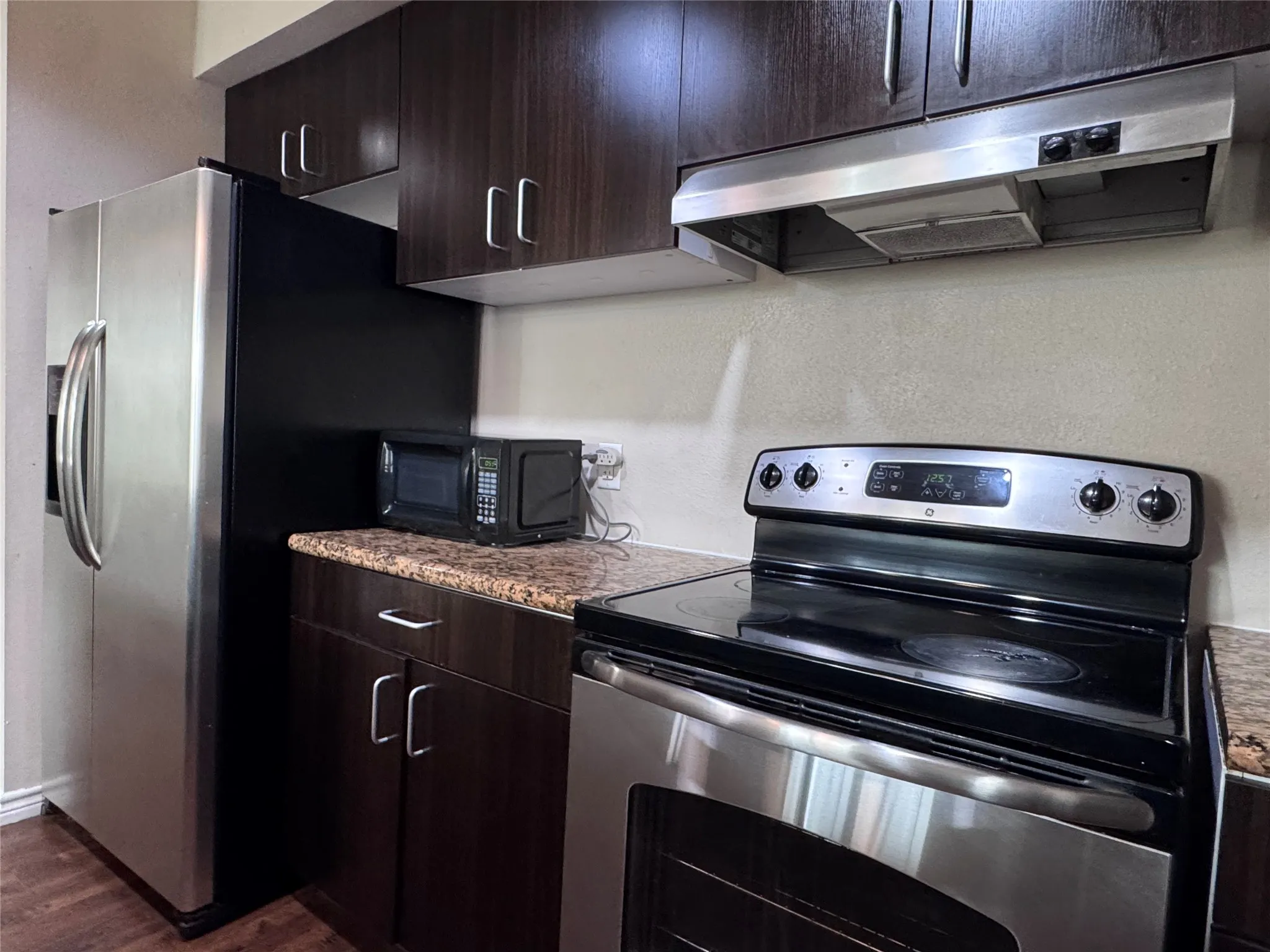 Stainless steel refrigerator, built-in stainless steel oven, dark wood-finish cabinetry, granite-finish countertops, and matching stainless steel range hood