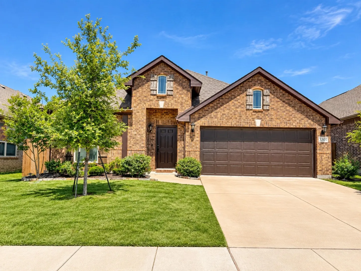 Brick facade with decorative window shutters, attached two-car garage, concrete driveway, and mature landscaping