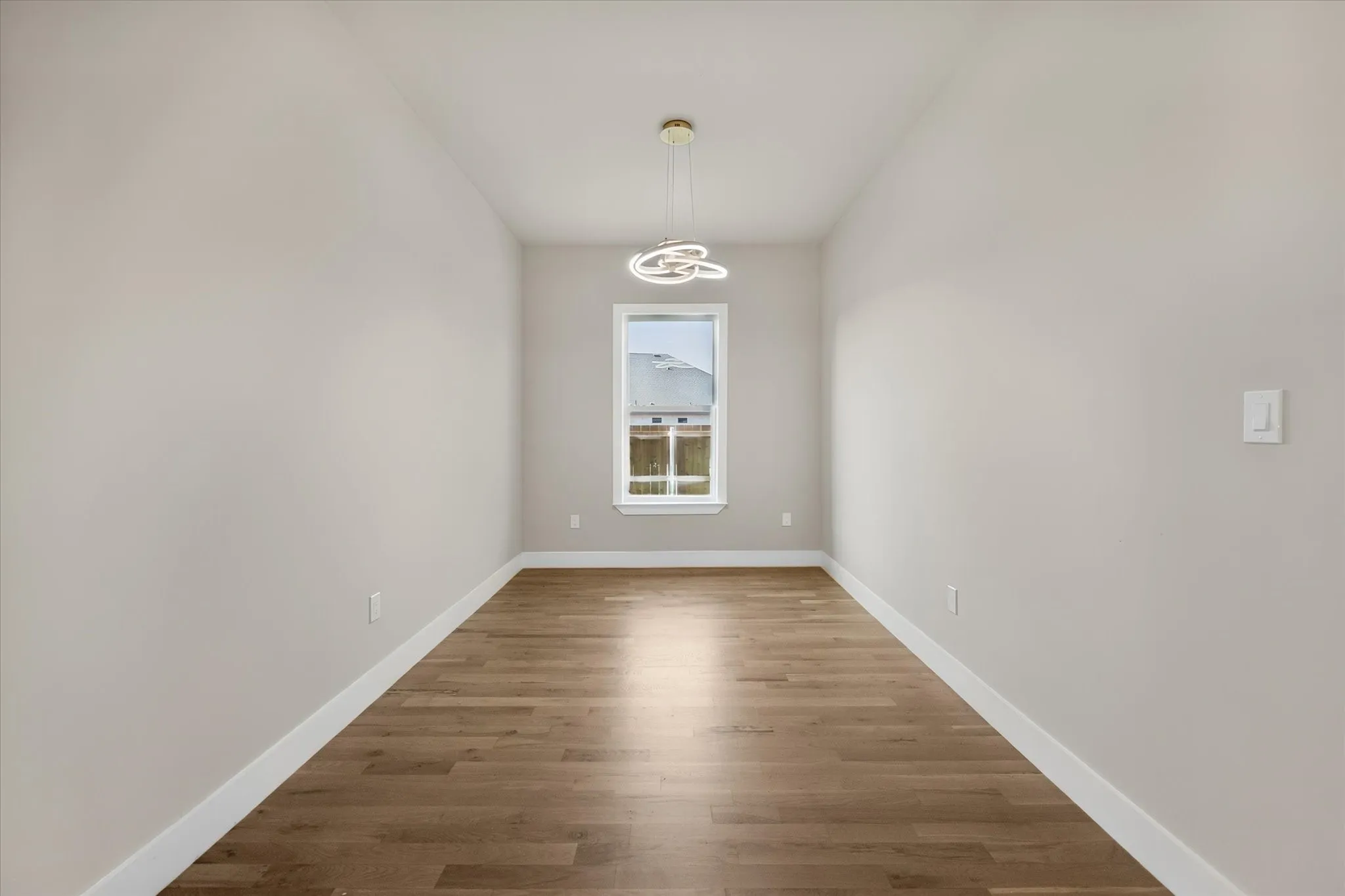 Room featuring light-toned walls, wood-finish flooring, a contemporary pendant light fixture, and a window with white trim