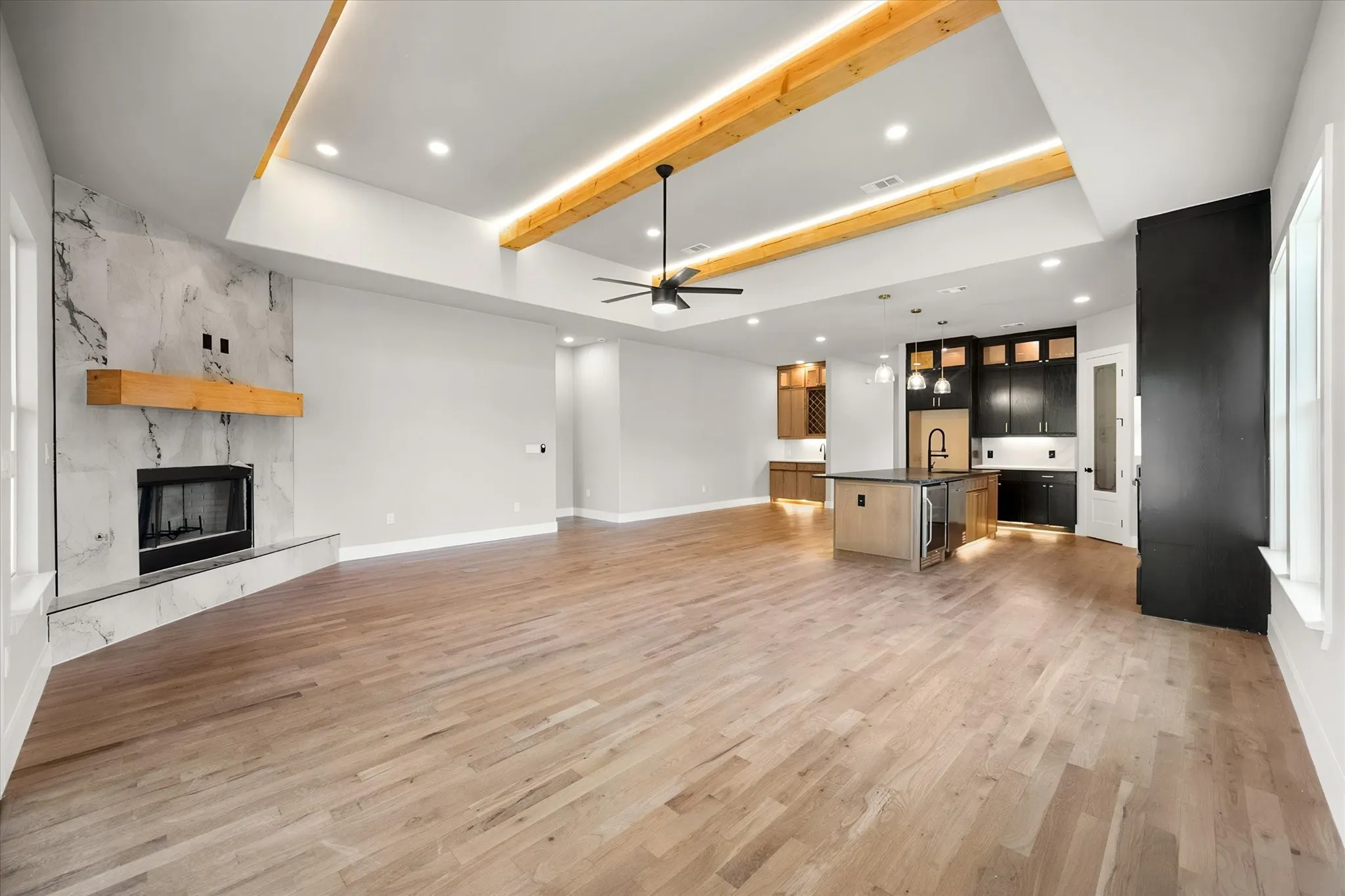 Light wood-finish flooring, vaulted ceiling with exposed wood beams and recessed lighting, tiled fireplace with wood mantel, kitchen island with built-in beverage refrigerator, and dark wood cabinetry