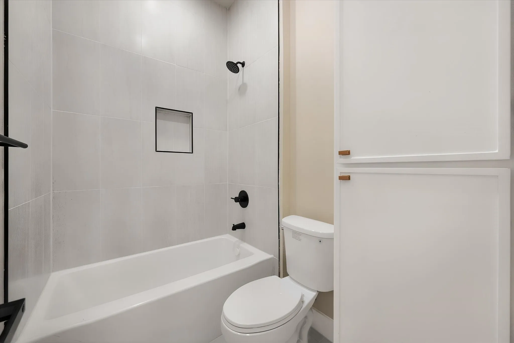 Bathtub with matte black fixtures, tiled surround, recessed shower niche, and adjacent built-in cabinetry