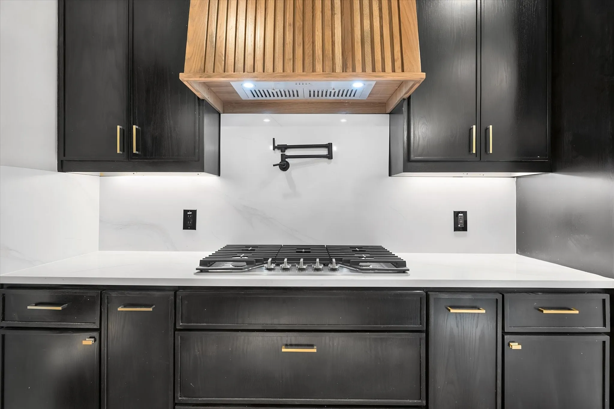 Custom wood range hood, black cabinetry with gold-tone hardware, white countertops, coordinating white backsplash, and gas cooktop